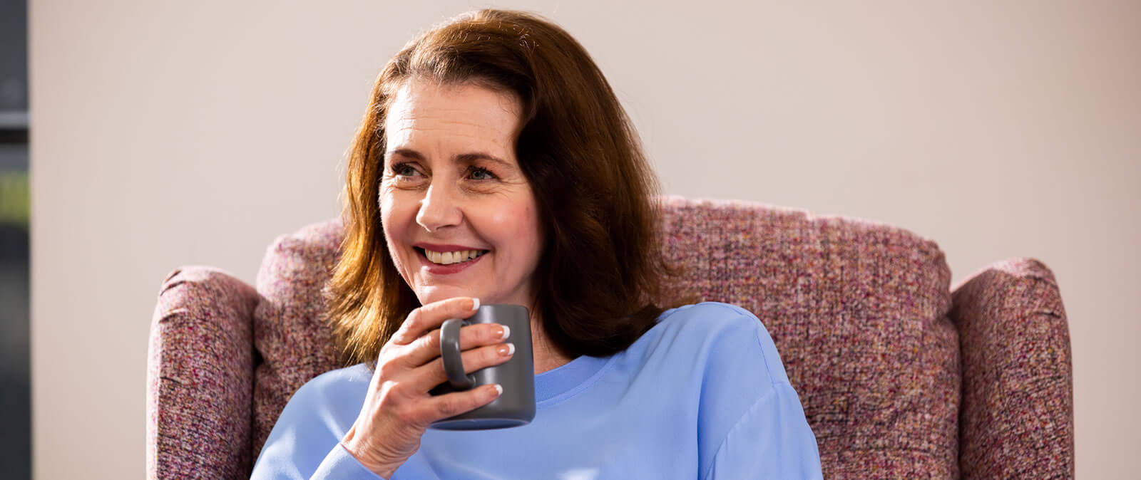 A middle-aged woman with brown hair, wearing a light blue jumper and holding a grey cup in her hand, with a friendly expression on her face.